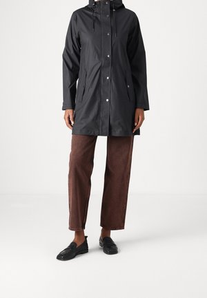 Person wearing a knee-length black hooded raincoat, brown wide-leg pants, and black loafers against a plain white background.