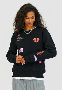 Čierna mikina s rôznymi farebnými nášivkami vrátane grafiky kvetu, srdca a homára, so zvinutými manžetami a lemom.