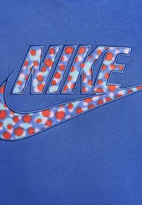 Blå Nike T-shirt med stort broderet "NIKE" logo med blomstermotiv i rød og blå, sat mod en ensfarvet blå baggrund.