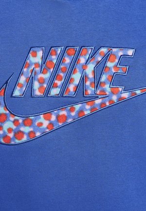 Blå Nike T-shirt med stort broderet "NIKE" logo med blomstermotiv i rød og blå, sat mod en ensfarvet blå baggrund.