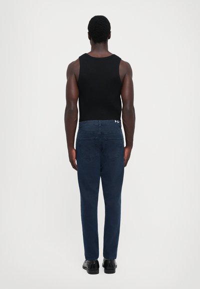Man standing facing away wearing a black tank top, dark blue jeans, and black shoes against a plain white background.