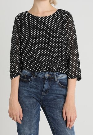 Schwarze durchsichtige Bluse mit weißen Punkten, runder Ausschnitt und dreiviertel Ärmel, kombiniert mit blauen Jeans.