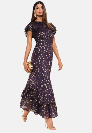 Mujer con vestido morado oscuro con manchas doradas, mangas y bajo con volantes, llevando un bolso de mano dorado y sandalias de tacón doradas.