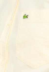 Τσέπη πουκαμίσου σε κρεμ χρώμα με μικρό πράσινο λογότυπο κροκόδειλου κεντημένο κοντά στην κορυφή.