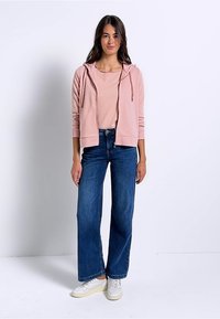 Felpa zip-up rosa chiaro sopra a una maglietta pastello, abbinata a jeans a gamba larga blu e sneaker bianche. Tessuto morbido e vestibilità casual.