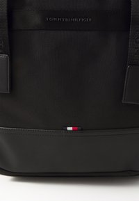 Czarny plecak z tkaniny o siateczkowej fakturze, z skórzanym dnem. Z przodu znajduje się logo "TOMMY HILFIGER" oraz mała naszywka w kolorach czerwonym, białym i niebieskim.