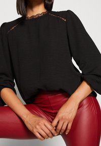 Blouse noire avec des détails en dentelle transparente au niveau de l'encolure et des manches bouffantes, associée à un pantalon rouge en similicuir moulant. Texture lisse dans l'ensemble.