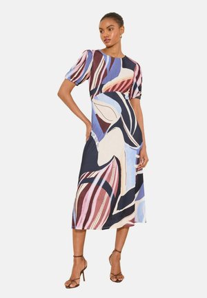 Žena nosiaca midi šaty s abstraktnými viacfarebnými vzormi, nafúknuté krátke rukávy a sandále na vysokom podpätku s remienkami, stojaca s jednou rukou na bokoch.