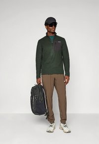 Žalias fliso megztinis su juoda kišenė su užtrauktuku ant krūtinės, rudos kelnės ir juodas kuprinė. Modelis dėvi kepurę ir akinius nuo saulės.