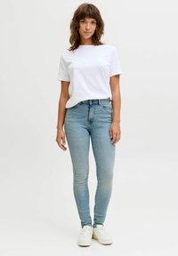 Biała koszulka z okrągłym dekoltem w zestawieniu z jasnoniebieskimi obcisłymi jeansami. Strój uzupełniają białe trampki. Gładki materiał i dopasowany krój.