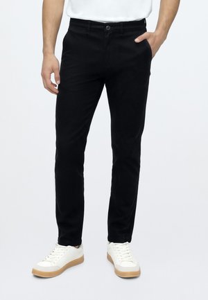 Homme portant un pantalon noir slim-fit avec bouton et fermeture éclair, une chemise blanche et des baskets blanches avec semelles en gomme, debout sur fond blanc.