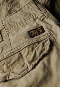 Close-up van de beige Superdry werkbroekzak met een zwart-gele stoffen merklap boven de stiksel van de zak genaaid.