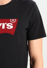 Svart t-shirt i bomull med en stor röd grafisk design med texten "LEVI'S®". Korta ärmar, rundad halsringning och en slät textur.