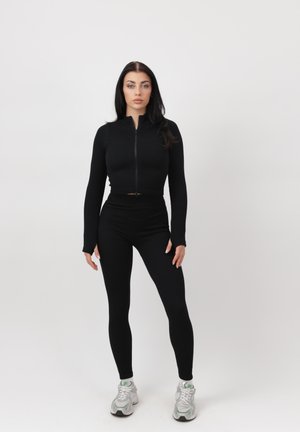 Kobieta stojąca prosto, ubrana w czarną bluzę na zamek z długim rękawem, czarne legginsy i białe trampki, na tle jednolitego jasnego tła.