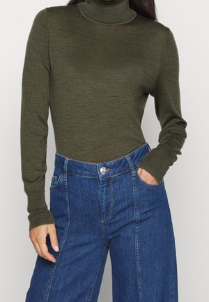 Personne portant un pull à col roulé vert olive rentré dans un jean en denim bleu taille haute, une main dans la poche.