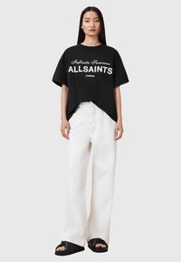 T-shirt oversize in cotone nero con la scritta "Infinite Summer ALLSAINTS LONDON" stampata in bianco, abbinata a pantaloni bianchi larghi e sandali neri.