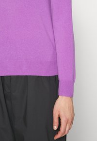 Lilla sweater med ribbet struktur, tilpassede manchetter og lige søm, parret med et sort plagg, der viser et nærbillede af ærmet.