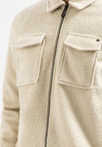 Beige, gestructureerde fleecejas met een rits aan de voorkant, kraag en twee borstzakken. Heeft contrasterende stiksels rond de zakken en een duurzame rits.