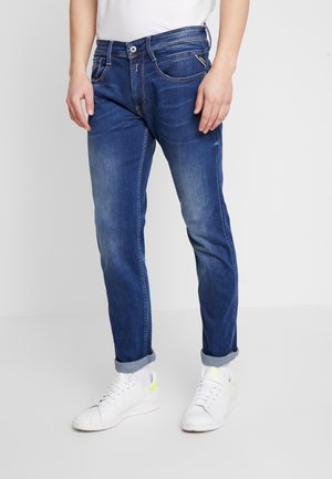 Man wearing blue rolled-up jeans and white sneakers, standing against a plain light background.