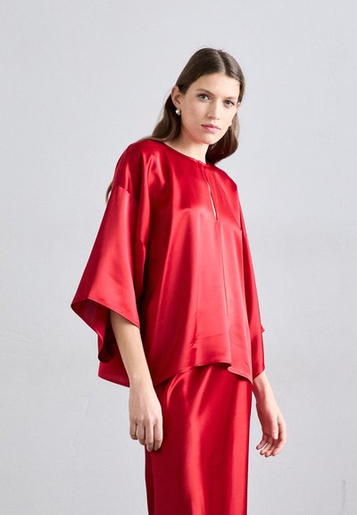 Blouse en satin rouge avec des manches larges, un col rond et un détail de fermeture éclair à l'avant. Le tissu a une texture lisse et brillante.