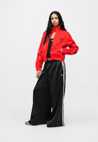 Rdeč sijoč tekaški pulover z visokim ovratnikom, črn logotip na prsih, kombiniran s črnimi hlačami širokih hlačnic z belimi stranskimi črtami.