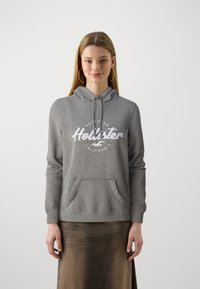 Gray hooded sweatshirt made of soft fabric with a front pocket. Features a white "Hollister" logo and text design on the chest.