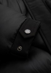 Giubbotto piumino nero con polsini abbottonati e bottoni a pressione circolari argentati, texture liscia e design trapuntato per il calore.