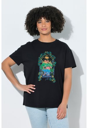 Kvinde med krøllet hår iført en sort T-shirt med et farverigt Capri-landskab og teksten "La Dolce Vita", kombineret med lyseblå jeans.