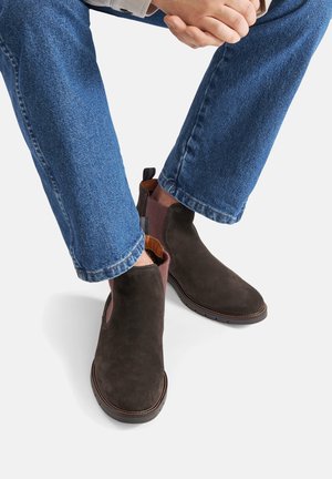 Braune Wildleder-Stiefeletten mit elastischen seitlichen Einsätzen in Burgunderrot, runder Zehenpartie und profillierter Gummisohle, kombiniert mit blauen Jeans.