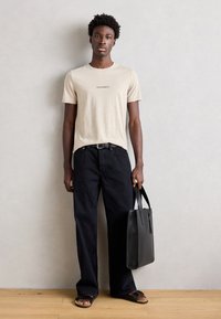 Tricou din bumbac bej cu textul "Arrivederci", pantaloni din denim negri cu picioare largi, geantă din piele neagră și sandale maro pe podea din lemn.