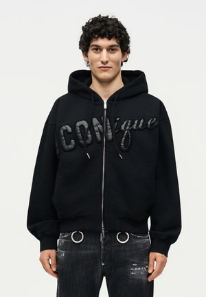 Νέος άνδρας με σγουρά μαλλιά που φοράει μαύρο φούτερ με φερμουάρ και υφασμένο κείμενο "Iconique" και φθαρτό μαύρο τζιν με μεταλλικούς κρίκους.