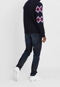 Marinblå tröja med diamantmönster i röd och vit på ärmarna, kombinerad med mörk denimjeans och vita sportskor.