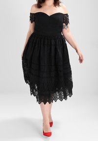 Černé šaty s odhalenými rameny, s texturovanou krajkovou pokládkou, přiléhavým vrškem a rozšířenou sukní. Mají zvlněný lem. Červené lodičky na vysokém podpatku.