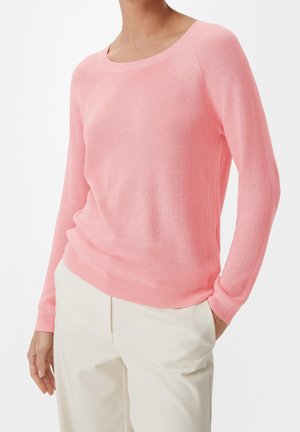 Femme portant un pull en maille rose clair à manches longues et un pantalon couleur crème, debout avec une main dans la poche devant un fond uni.