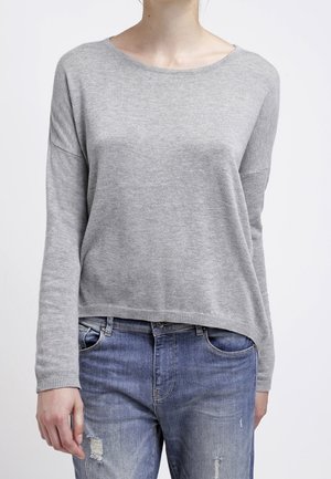 Person iført en lysegrå, langærmet sweater og slidte blå jeans, stående mod en ensfarvet hvid baggrund.