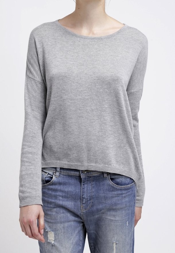 Personne portant un pull gris clair à manches longues et un jean bleu déchiré, debout sur un fond blanc uni.