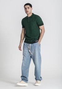 Polo de punto verde con mangas cortas, que cuenta con un bolsillo y ribetes a rayas. Combinado con unos vaqueros holgados de color azul claro y zapatillas crema.