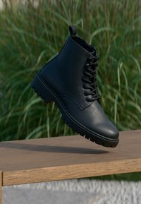 Stivale alla caviglia in pelle nera con lacci frontali, suola resistente e linguetta. Texture liscia con cuciture minime e silhouette classica.