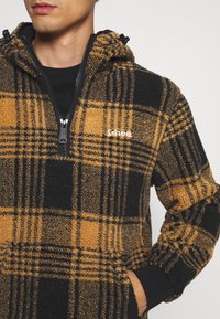 Sweat à capuche en polaire à carreaux marron et noir avec une fermeture éclair à moitié, cordon de serrage ajustable et poches avant. Nom de la marque discrètement affiché.