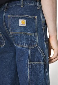 Pantalones de mezclilla en azul oscuro con costuras visibles. Incluye varios bolsillos y una etiqueta cuadrada con un logo amarillo en la parte trasera.