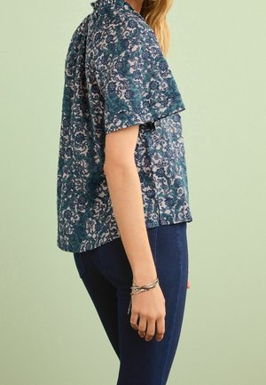 Blusa a maniche corte blu con motivo, dotata di un colletto alto, caratterizzata da disegni intricati e una vestibilità ampia; abbinata a jeans blu scuro.