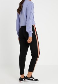 Kobieta w lawendowym swetrze z teksturowanymi rękawami oraz czarnych spodniach z beżowymi i czerwonymi paskami po bokach, do których założyła czarne trampki.