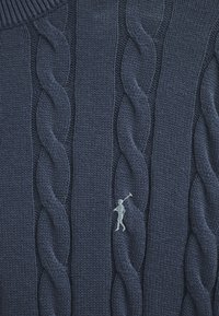 Mörkblå stickad tröja med ett strukturerat flätmönster och en liten vit broderad figur som håller i en golfklubba på framsidan.