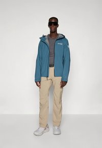 Casaco impermeável azul claro com capuz, forro interno cinza, combinado com camisa cinza e calças bege. Modelo usa óculos de sol e ténis.