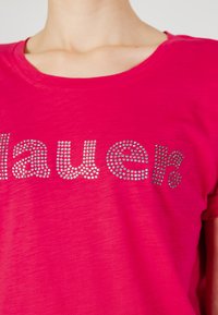 T-shirt rosa brillante con maniche corte, presenta un design testurizzato di strass che compongono la scritta "laue". Vestibilità leggermente ampia e scollatura a girocollo.