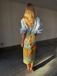 A woman stands in a bright room, wearing a light blue blouse and a colorful patterned skirt, with sunlight casting shadows on the floor.