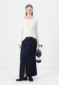 Maglietta bianca a coste con maniche lunghe abbinata a una gonna maxi in denim scuro con spacco anteriore. Scarpe nere e una piccola borsa nera completano l'outfit.