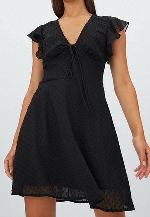 Vrouw draagt een zwarte jurk van gestructureerde stof met vlindermouwen en een V-hals met veterdetail, staand tegen een effen lichte achtergrond.