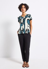 Blusa de patrón multicolor en teal, blanco y negro, combinada con pantalones negros ajustados y sandalias negras con tiras.