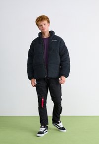 Zwarte fleece jack met een rits aan de voorkant, oversized ontwerp, geribbeld kraag, gedragen over een paarse shirt, gecombineerd met zwarte cargo pants en sneakers.